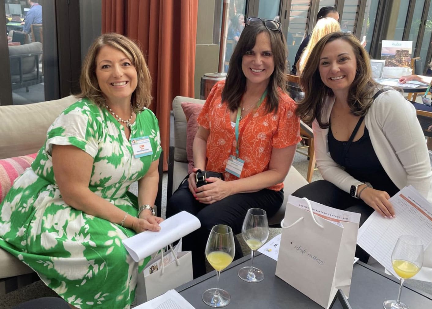 Three women are seated together indoors, smiling at the camera. Papers, glasses, and a bag are on the table in front of them. People are visible in the background.