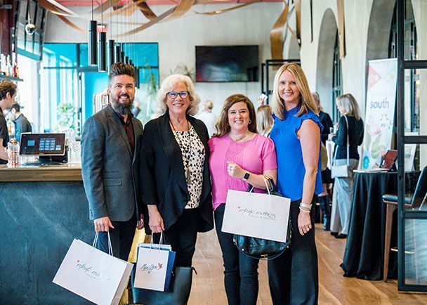 Four people standing in a restaurant, holding Retreats Resources shopping bags, and smiling at the camera. Tables and other people are in the background.