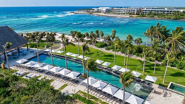 Conrad Tulum beachfront resort with a row of poolside lounge chairs and umbrellas, palm trees, and turquoise ocean water in the background under a clear sky.