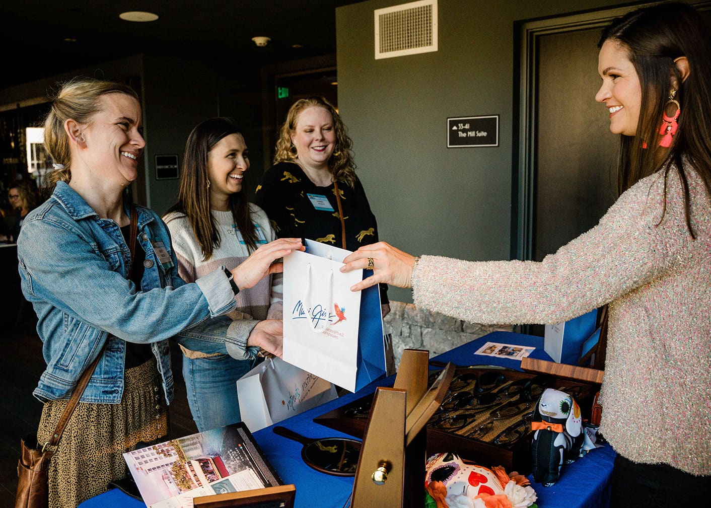 Four women at a booth during a Retreats Resources Roadshow event. Three are smiling and one receives a Maui Jim gift bag from the woman across the table.
