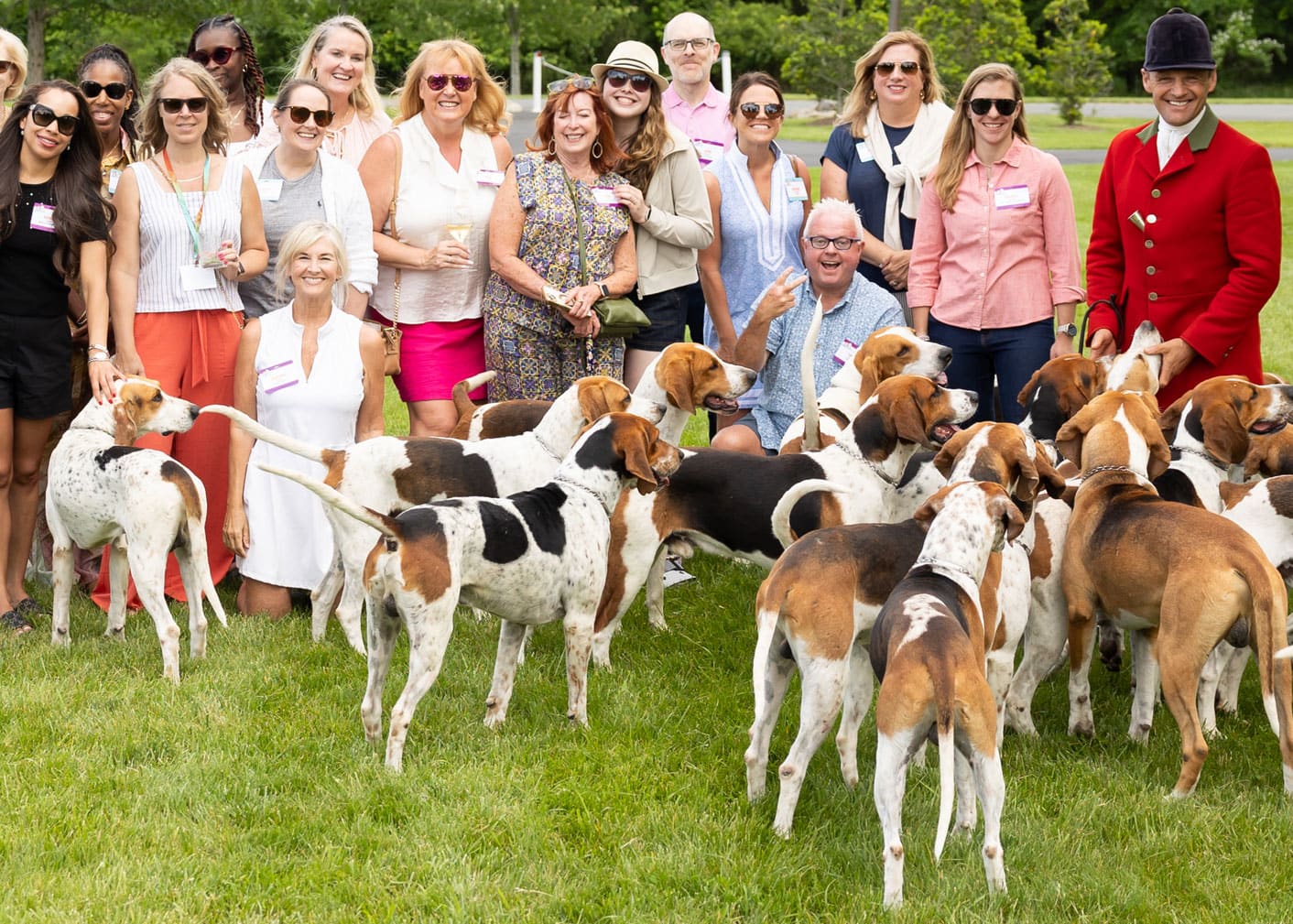 A group of people posing outdoors on grass with several hounds in front of them; trees and greenery are in the background.
