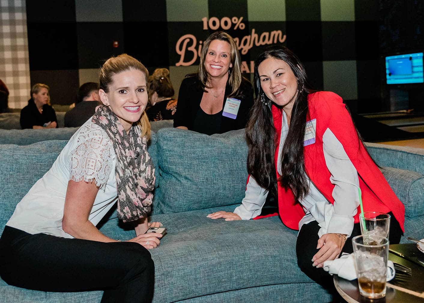 Three women sit and smile on a blue couch at an indoor event; drinks are on the table in front of them, and a sign in the background reads “100% Birmingham”.