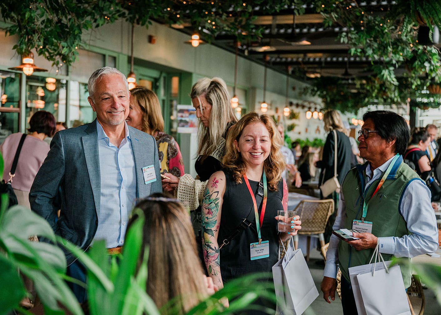 A group of people wearing name badges and event lanyards converse and smile at an indoor gathering with greenery and ambient lighting.