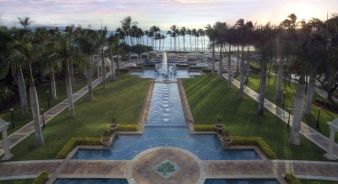 A landscaped resort garden at Grand Wailea, A Waldorf Astoria Resort, features palm trees, fountains, pools, and walkways leading toward the ocean, with cabanas and lounge chairs visible near the water.