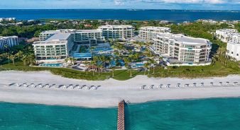 Aerial view of The St. Regis Longboat Key Resort - a large beachfront resort with multiple buildings, pools, palm trees, and beach chairs lined up near turquoise water under a partly cloudy sky.