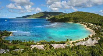 Aerial view of a tropical bay with turquoise water, green hills, a sandy beach, and scattered huts of Rosewood Little Dix Bay, with a sailboat in the distance under a partly cloudy sky.