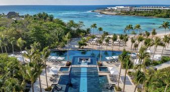 Aerial view of Conrad Tulum Riviera Maya, a luxurious beachfront resort with palm trees, an infinity pool, lounge chairs, and a sandy beach overlooking turquoise ocean waters.