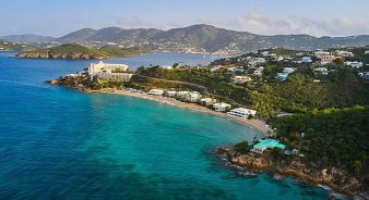 Aerial view of a coastal area with turquoise water, sandy beach, residential buildings, and The Westin St Thomas Beach Resort & Spa at the end of a peninsula, with green hills in the background.