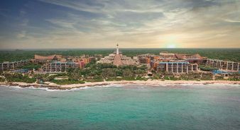 Aerial view of Hotel Xcaret Arte, a large beachfront resort complex surrounded by greenery, with buildings facing the ocean and the sun setting in the background.