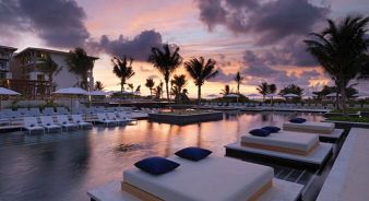 Outdoor pool area at a resort with poolside beds and lounge chairs, palm trees, and buildings, at sunset with a colorful sky.