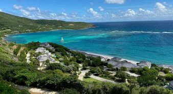 Aerial view of a tropical coastline with turquoise water, a sailboat, green hills, and Mandarin Oriental, Canouan Resort nestled among lush vegetation.