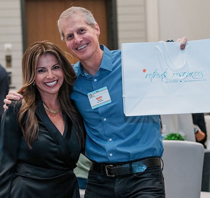 Two people smiling, with the man holding up a white bag labeled "Retreats Resources." Both wear professional attire, and the man has a name tag, Scott.