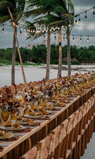 An elegant outdoor dining setup features a long table with decorative centerpieces and string lights above, set against a backdrop of palm trees and a beach.