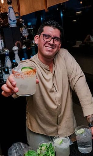 A bartender smiles while reaching out to offer a cocktail with lime garnish. He is behind the bar counter and surrounded by bottles in the background.
