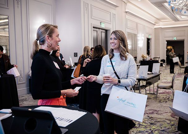 Two women exchanging business cards at a Retreats Resources roadshow event. Other attendees are in the background, and one woman holds a shopping bag.