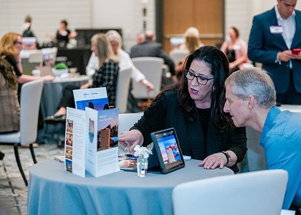 Two people at a table discussing materials, with a tablet and brochures in front of them. Several other people are in the background in a roadshow setting.